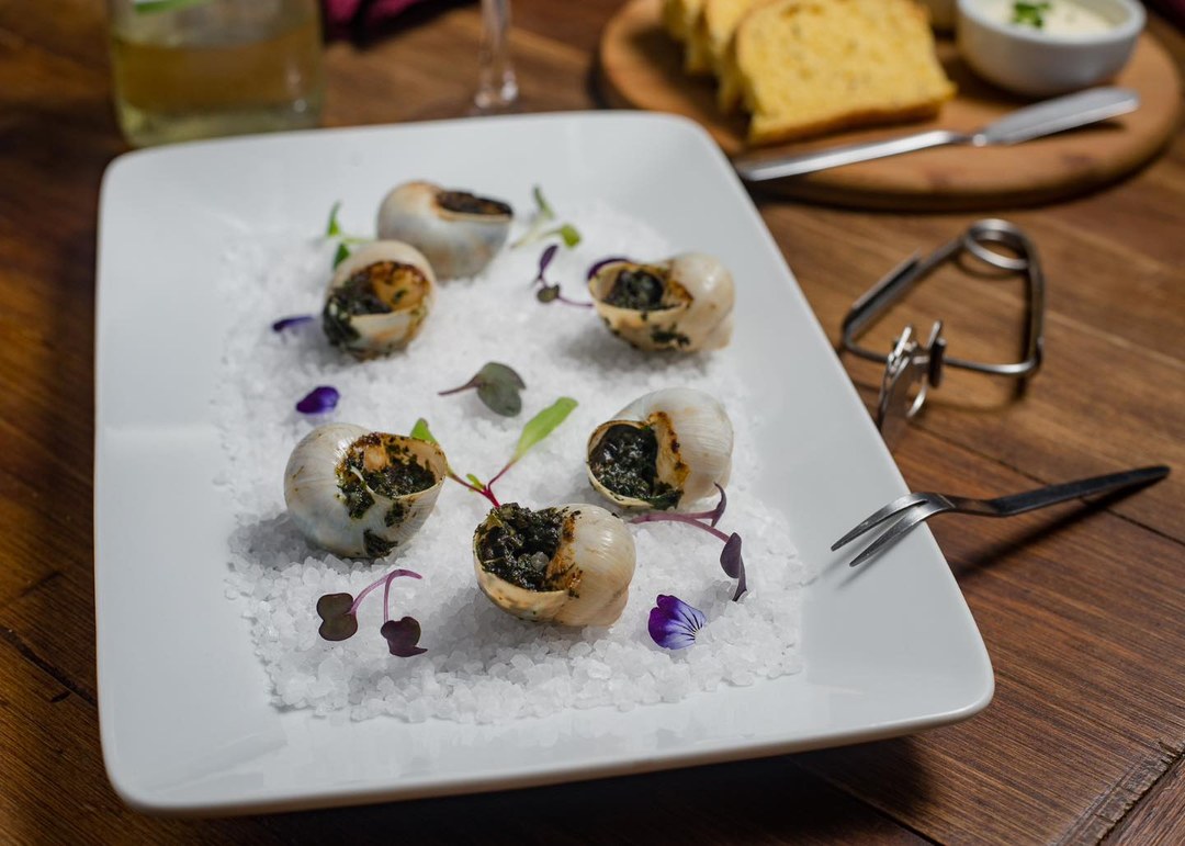 Conchas de escargot assadas sobre um leito de sal, em prato quadrado branco.