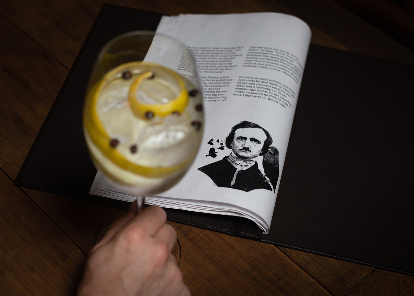 Um drinque com casca de limão apoiado sobre um livro aberto na última estrofe de O Corvo, ao lado de uma gravura de Edgar Allan Poe com um corvo no ombro.
